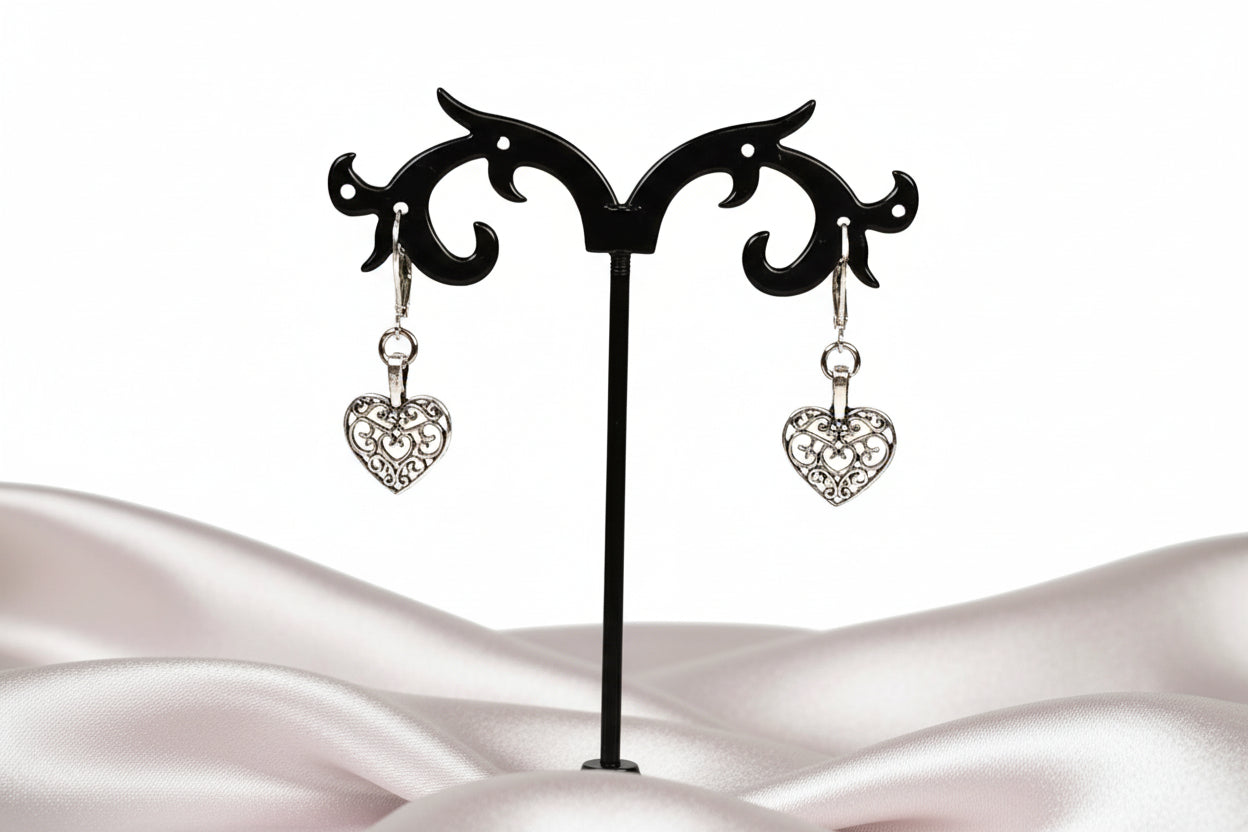 Dangling silver heart-shaped earrings displayed on a black stand, with the earrings featuring a clasp closure.