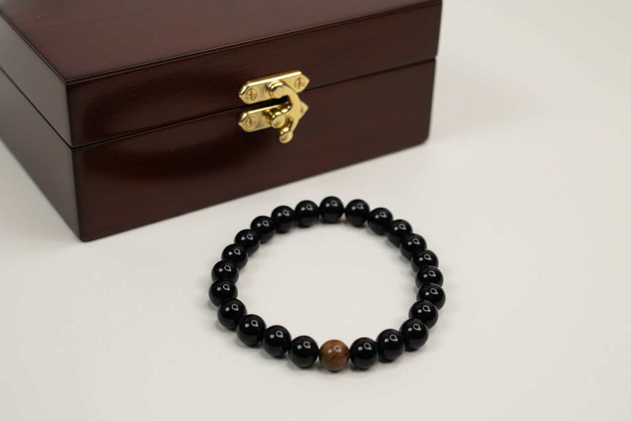 Black beaded bracelet with a brown stone on a white surface, next to a brown jewelry box.