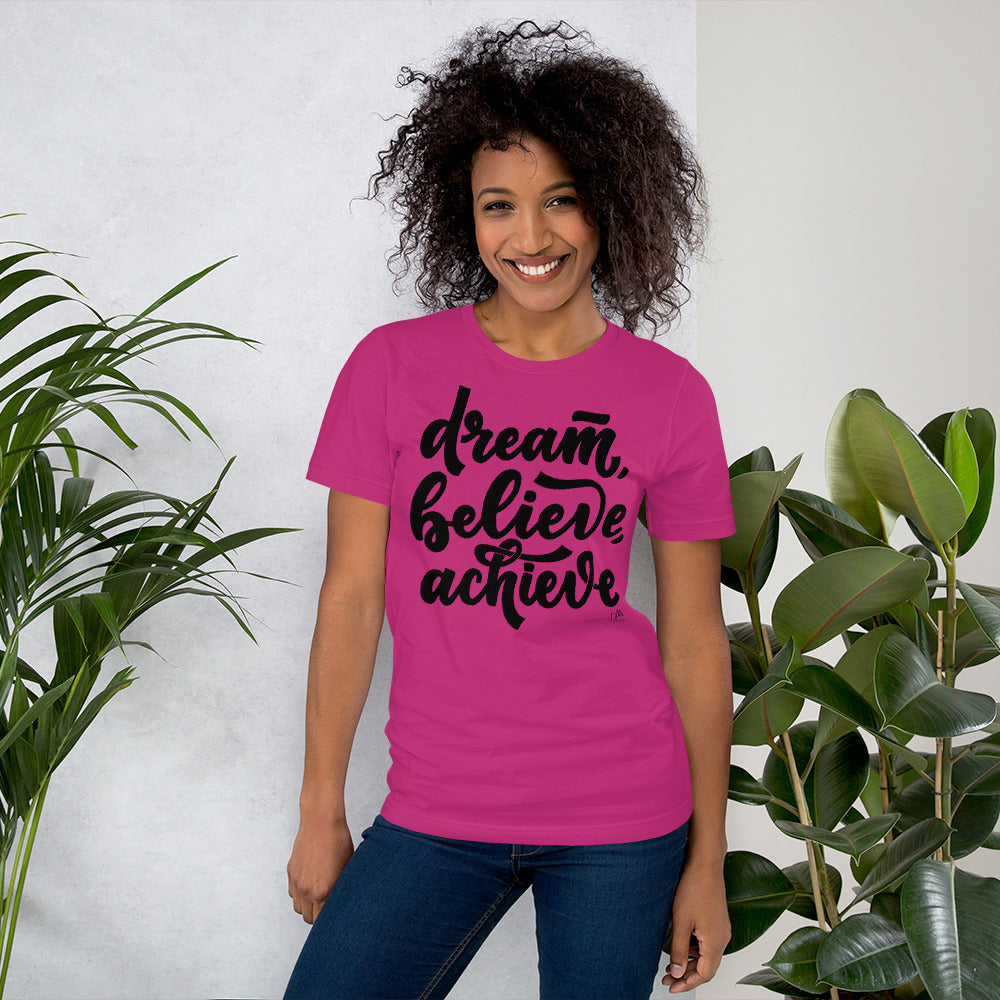 Woman wearing a pink t-shirt with 'dream, believe, achieve' text, standing between two plants.