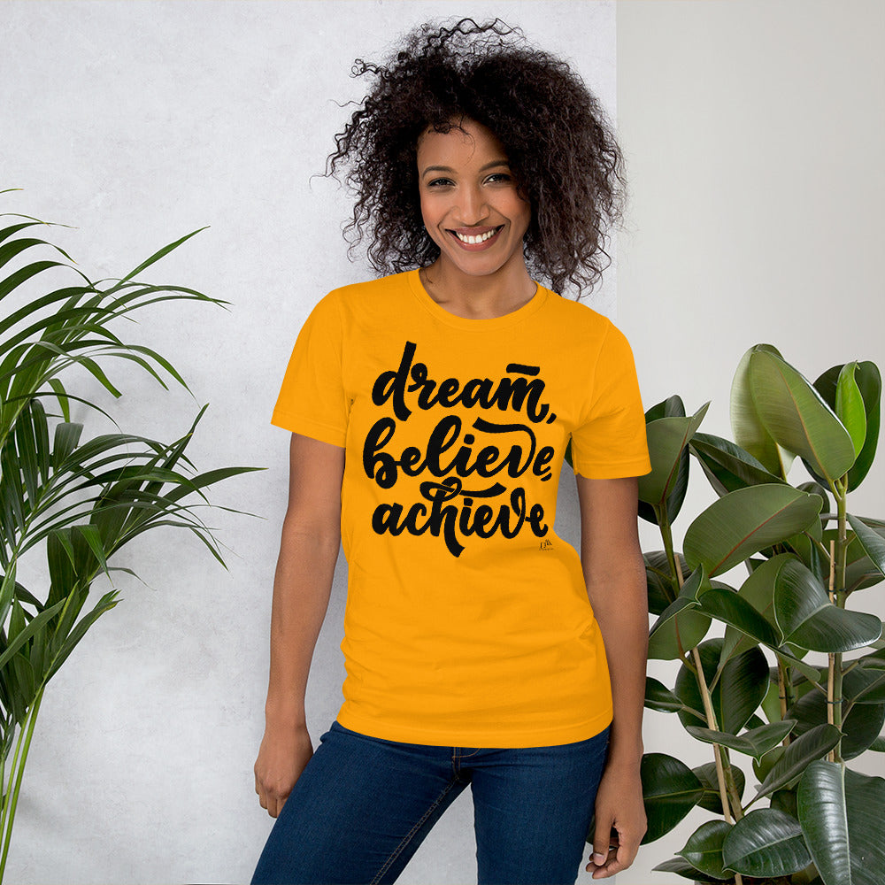 Woman wearing a yellow t-shirt with 'dream, believe, achieve' text, standing between two plants.