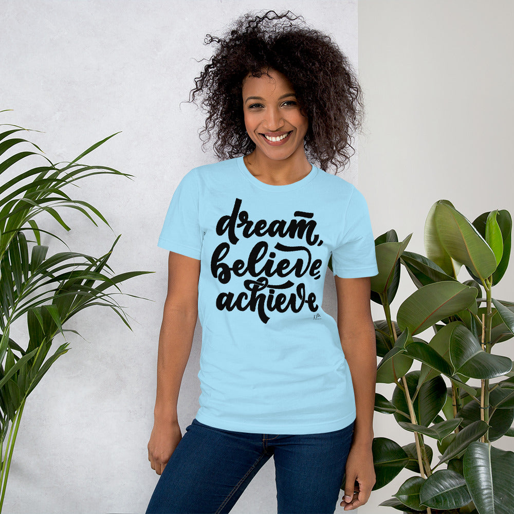 Woman wearing a light blue t-shirt with 'dream, believe, achieve' text, standing between two plants.