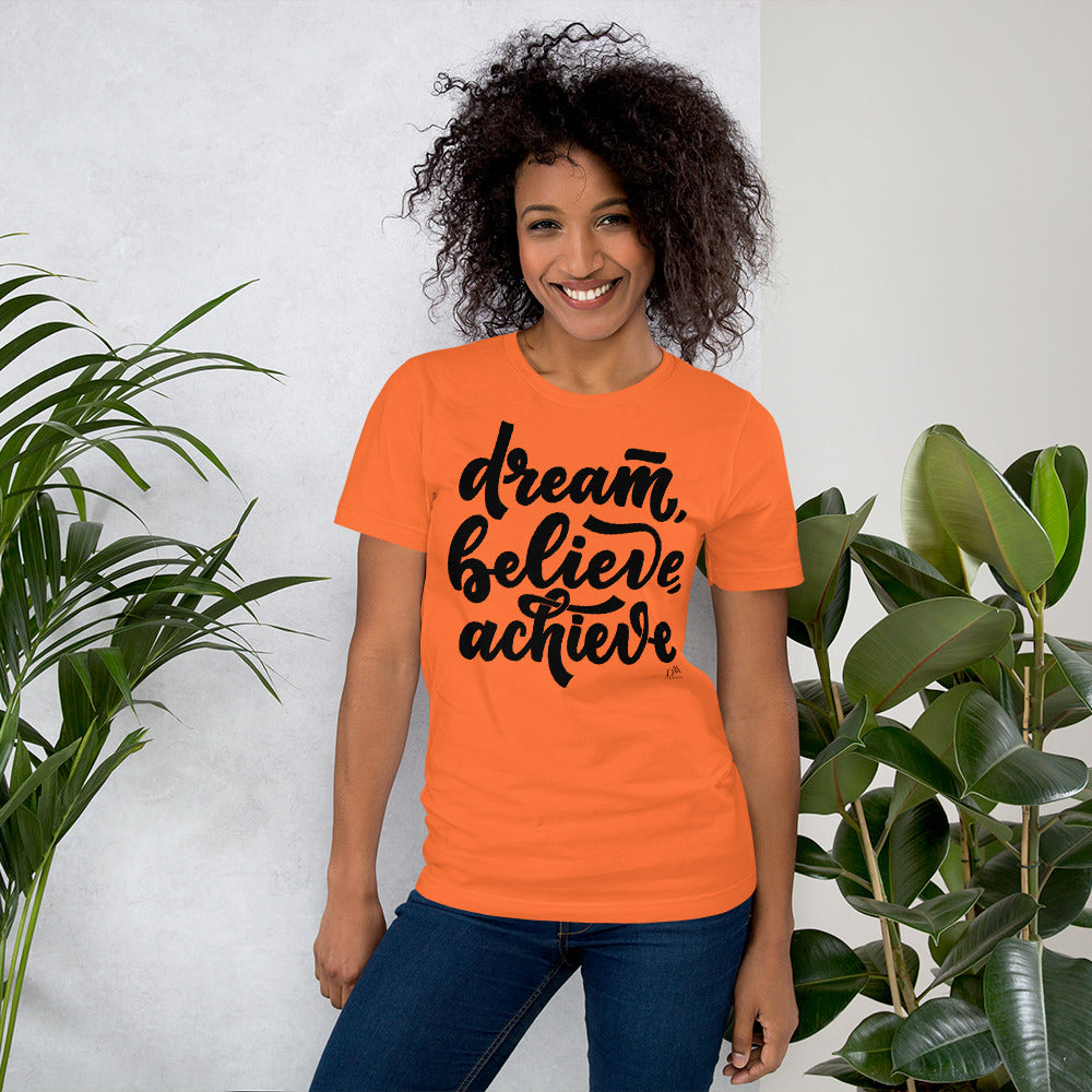Woman wearing an orange t-shirt with 'dream, believe, achieve' text, standing between two plants.