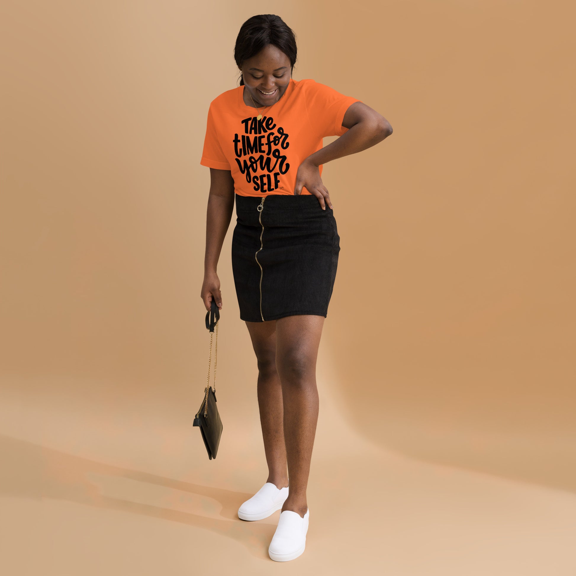 Woman wearing an orange t-shirt with text, black skirt, and white shoes on a beige background