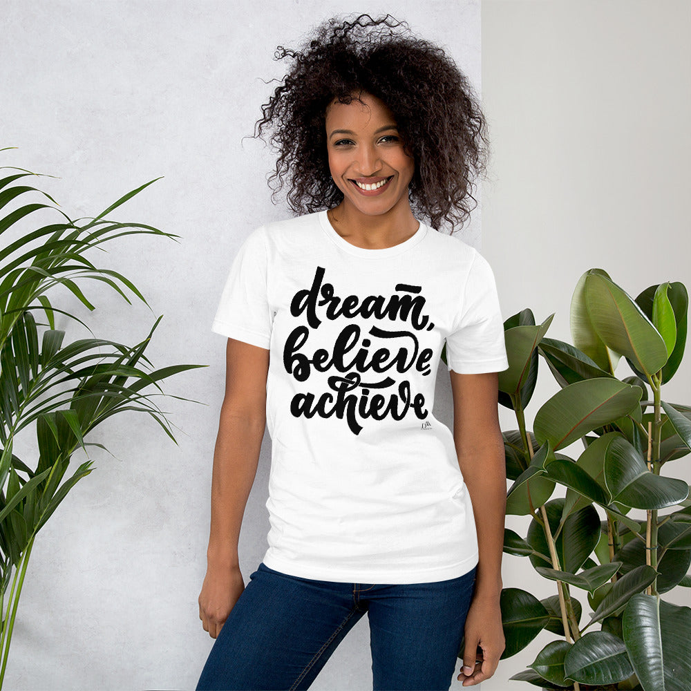 Woman wearing a white t-shirt with 'dream, believe, achieve' text, standing between two plants.